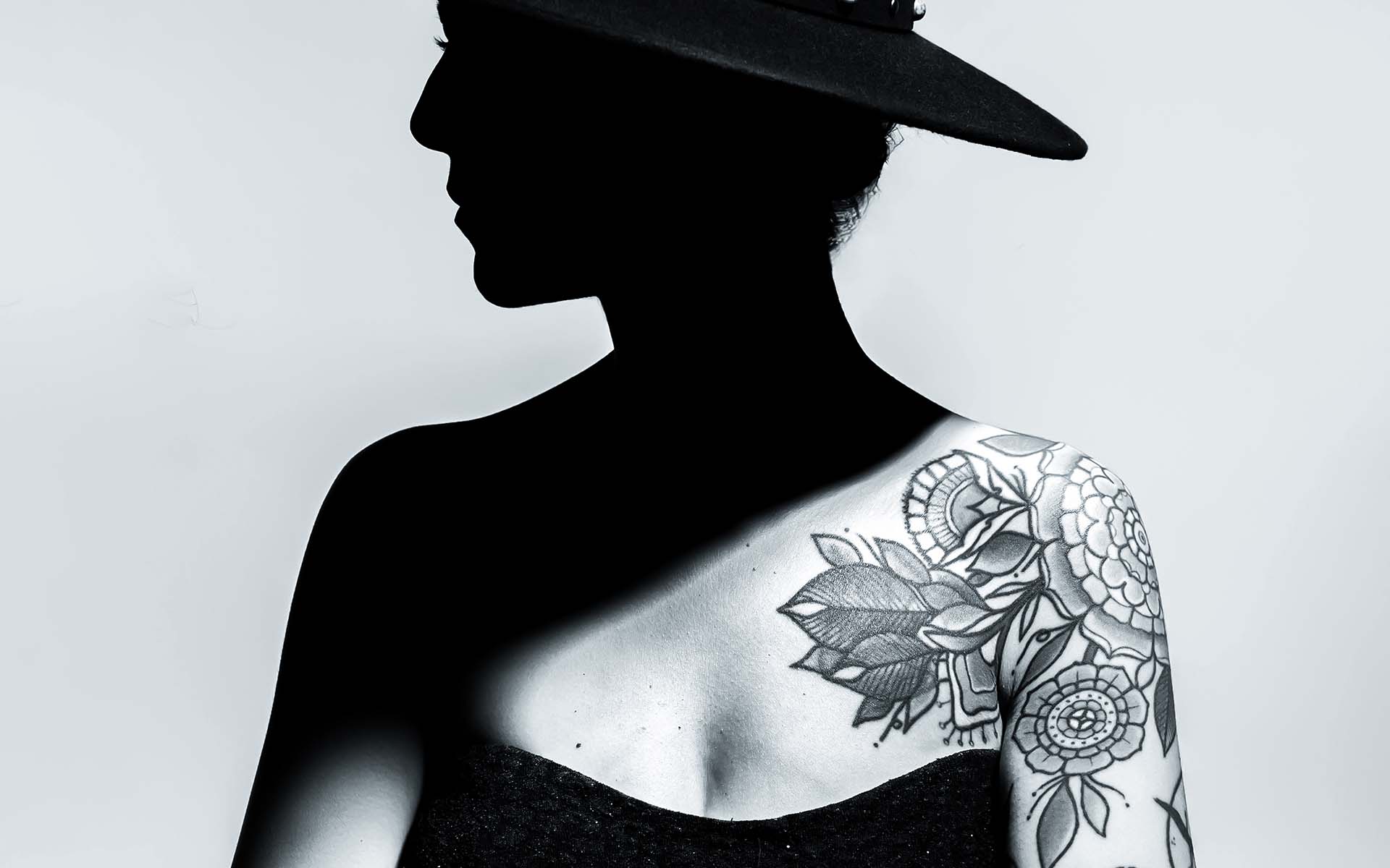 Silhouette of a woman wearing a wide-brimmed hat, showcasing intricate floral tattoos on her exposed shoulder and back.